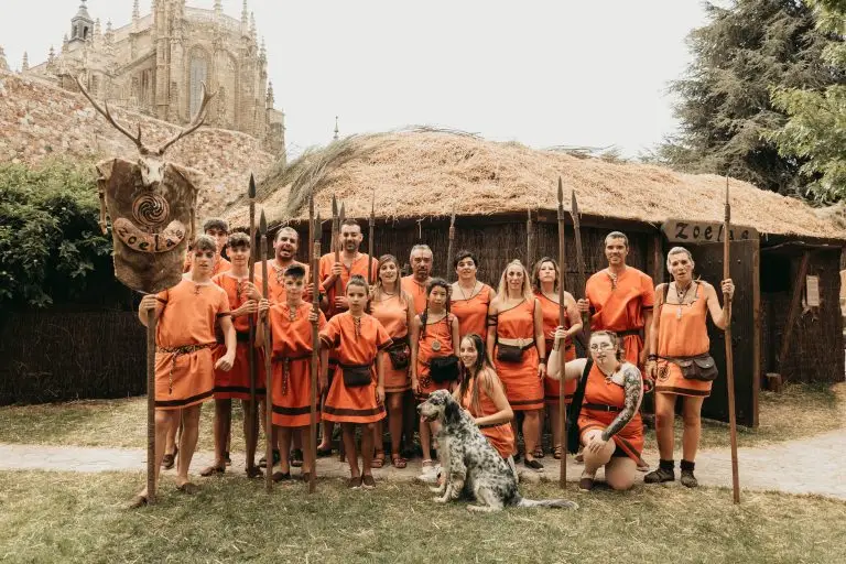 Foto de grupo de la tribu astur Zoelas con túnicas naranjas y lanzas frente a su palloza en el poblado, con la Catedral de Astorga al fondo, Fiesta de Astures y Romanos