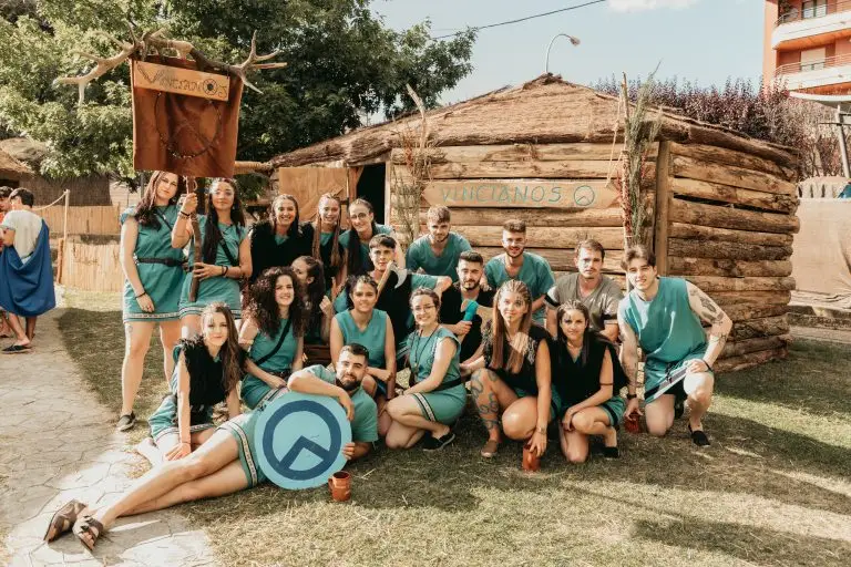 Foto grupal de la tribu astur Vincianos con indumentaria verde azulada, escudo tribal y cuernos de ciervo frente a su palloza de troncos en el poblado astur de Astorga
