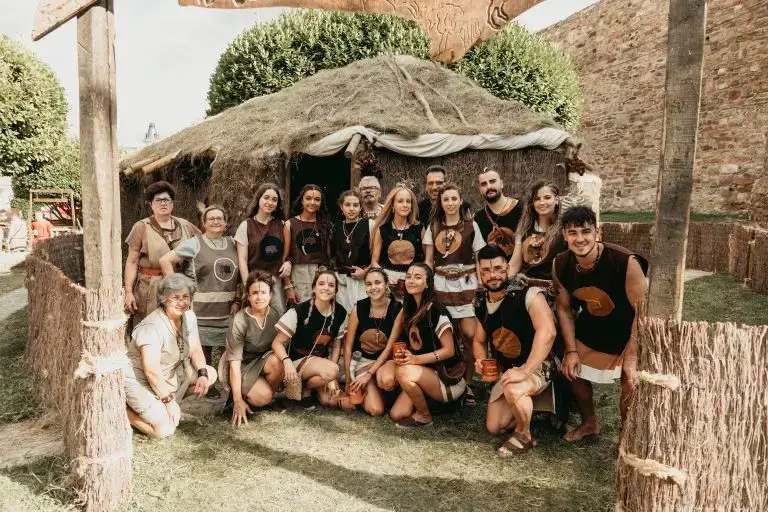 Foto grupal de la tribu astur Orniacos con túnicas marrones y el emblema del jabalí bordado, frente a su palloza junto a la muralla romana de Astorga