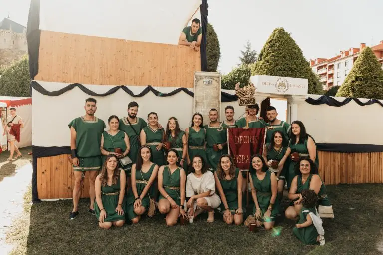 Foto grupal de Decivs Hispanvs con túnicas verdes, estandarte y entrada de domus con águila romana y sello SPQR en el campamento romano de Astorga