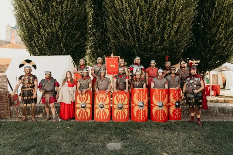Foto grupal de la Legio VI Victrix con armaduras romanas completas, escudos rojos con águilas doradas y estandarte legionario en el campamento romano de AstorgaTítulo: Legio VI Victrix - Legión romana de Astu
